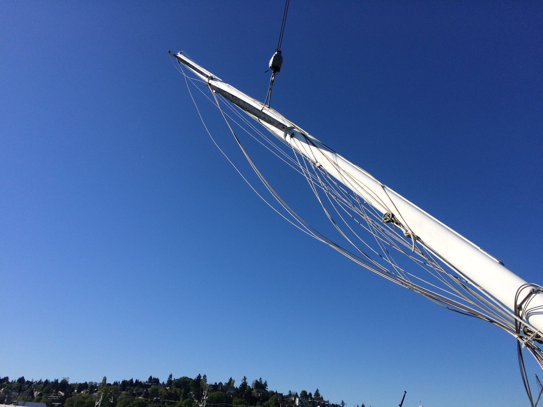 The graceful boom and rigging of the classic 1946 Merry Maiden silhouetted against a crystalline Puget Sound sky, her varnished spar gleaming in brilliant sunlight with evergreen-lined shores distant below.