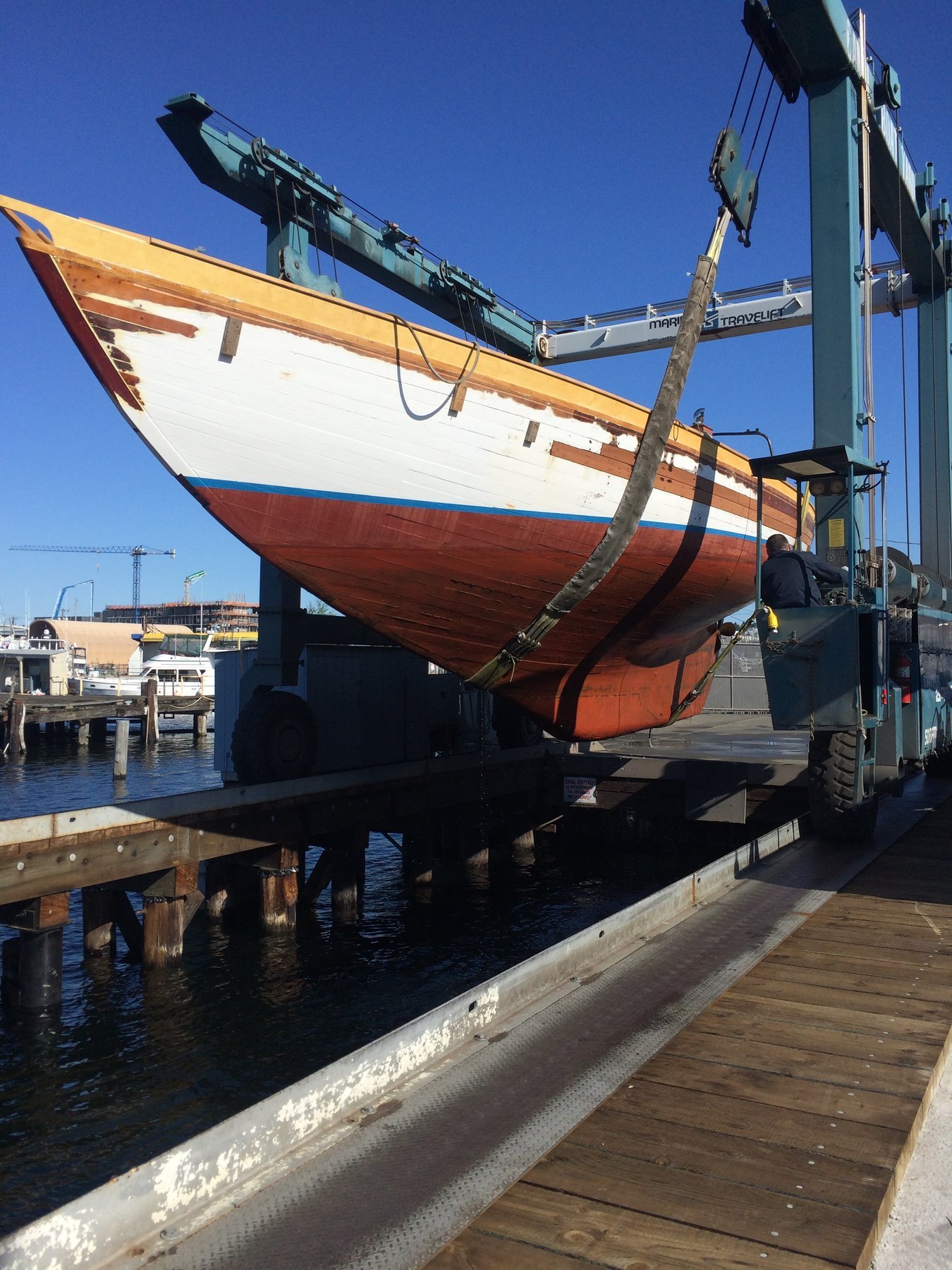 The classic 1946 Philip Rhodes-designed Merry Maiden sits cradled in a Puget Sound boatyard, her distinctive red hull and yellow boot stripe gleaming against the clear blue sky as she awaits maintenance or restoration work.