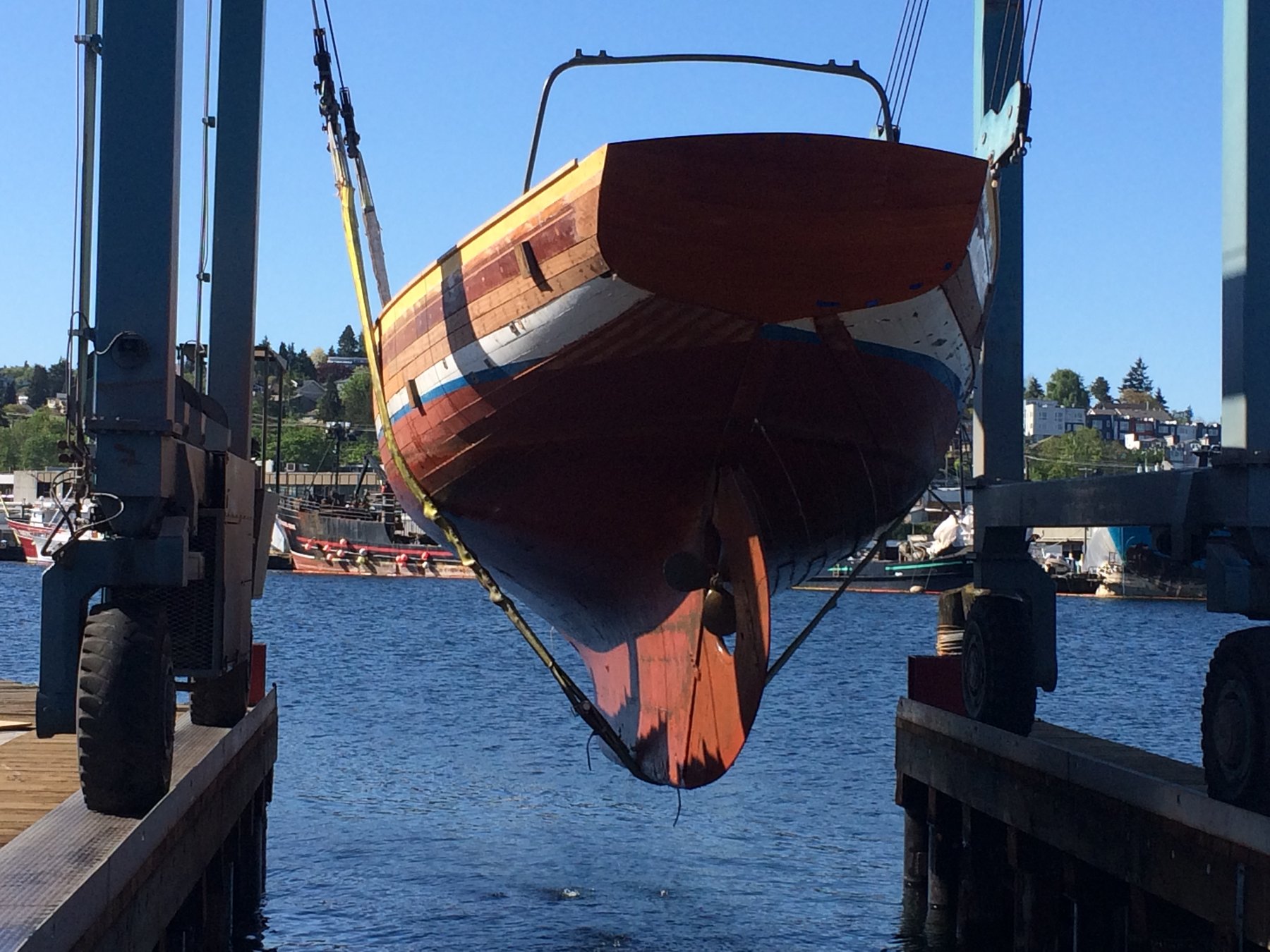 The classic 1946 Philip Rhodes-designed Merry Maiden hangs suspended in the boat hoist at Puget Sound, her wooden hull and colorful stripe pattern gleaming in the spring sunlight as she transitions between water and land.