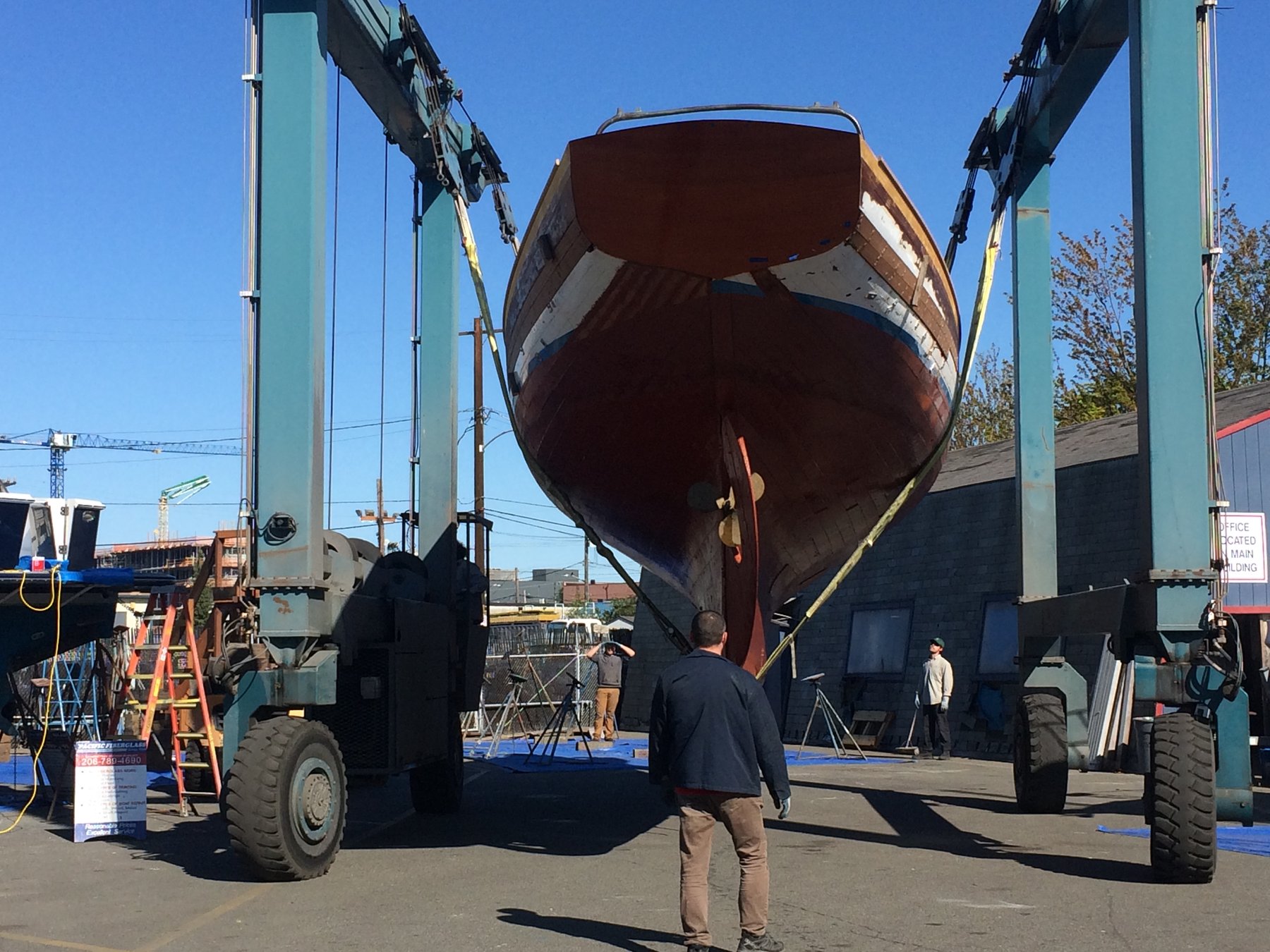 The Merry Maiden, a 1946 Philip Rhodes classic, is carefully lifted from the water in a Puget Sound boatyard, suspended between the towering green posts of a heavy-duty travel lift.