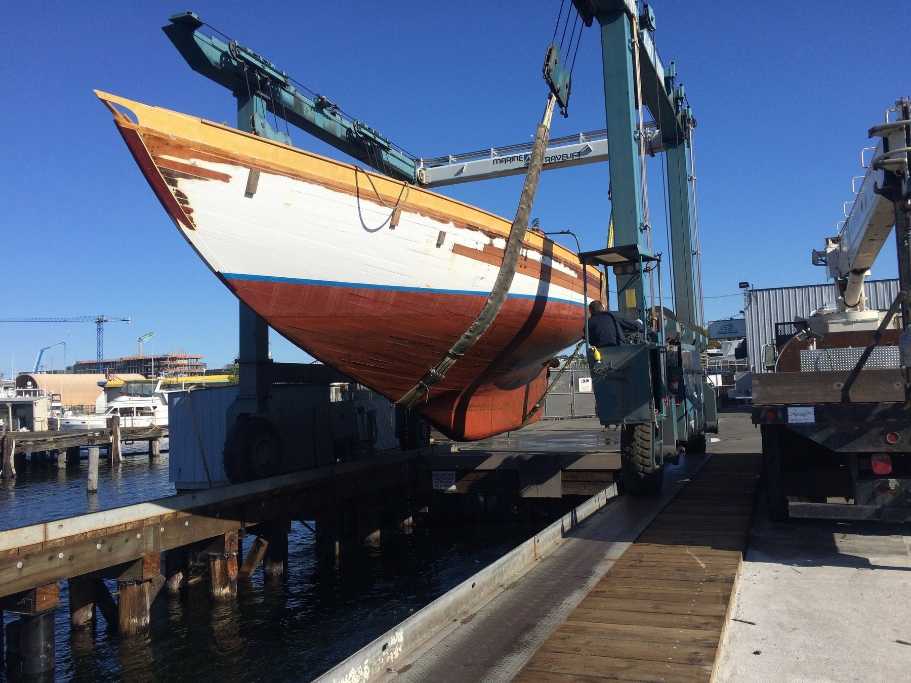 The classic 1946 Philip Rhodes-designed Merry Maiden suspended in a travel lift at a Puget Sound boatyard, her iconic red hull and cream topsides gleaming against the clear blue sky.