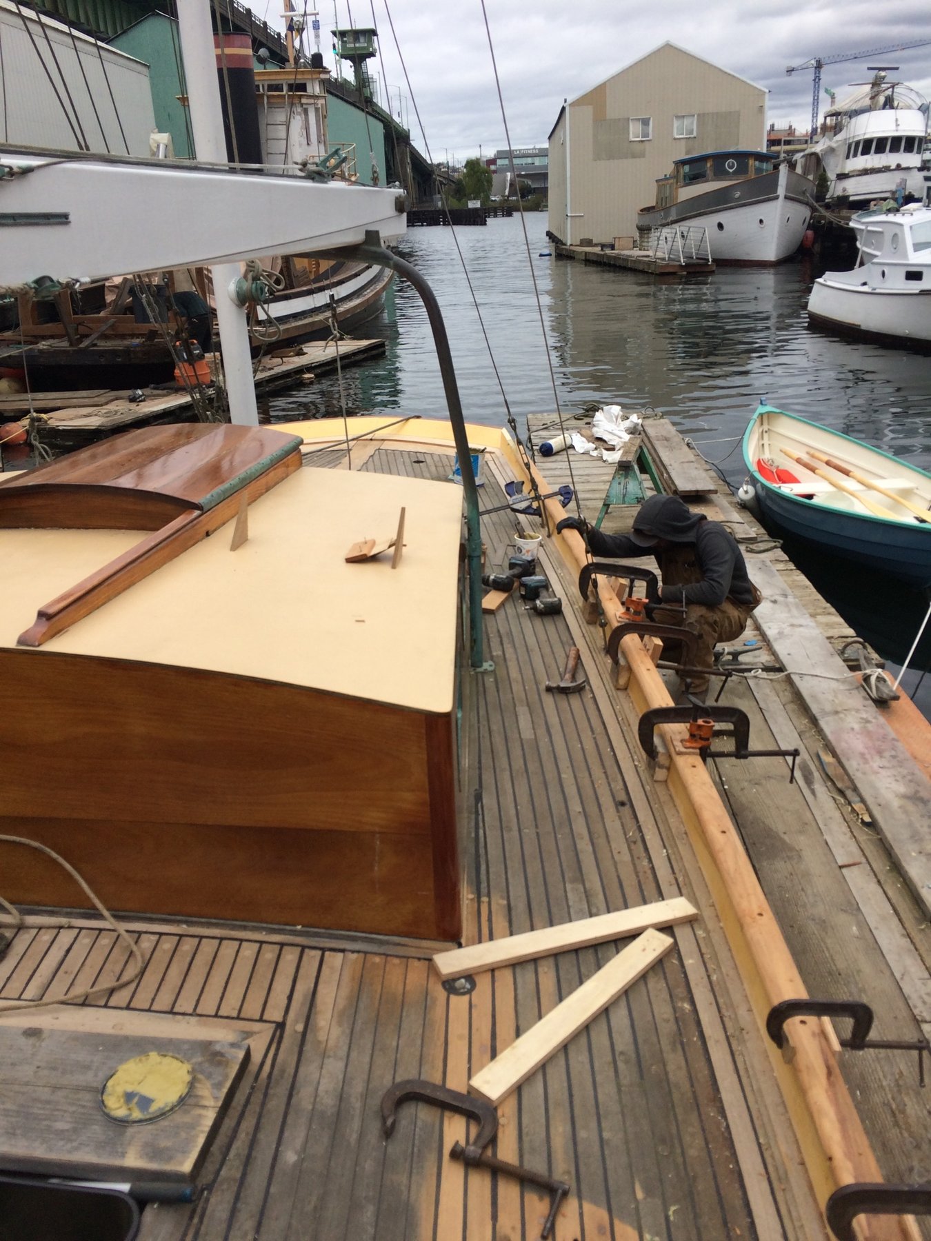 Merry Maiden's teak deck gleams in soft spring light, her classic lines and traditional wooden fittings a testament to 1946 craftsmanship, moored among working boats in a historic maritime basin.