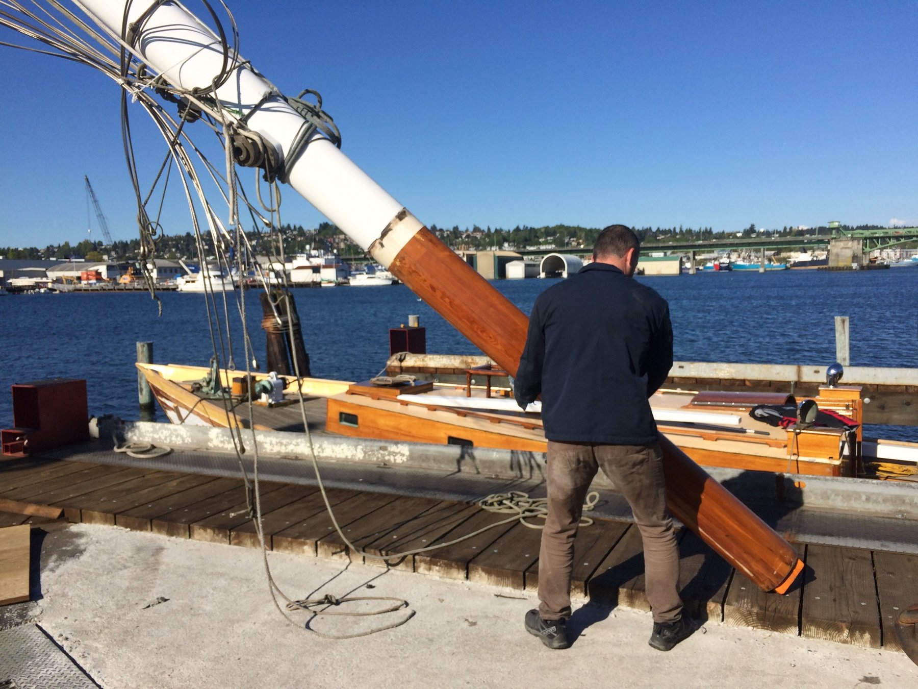 A crew member guides the Merry Maiden's wooden mast during dockside work, with the classic 1946 yacht's distinctive cabin sides gleaming in the spring sunshine at what appears to be a Pacific Northwest maritime facility.
