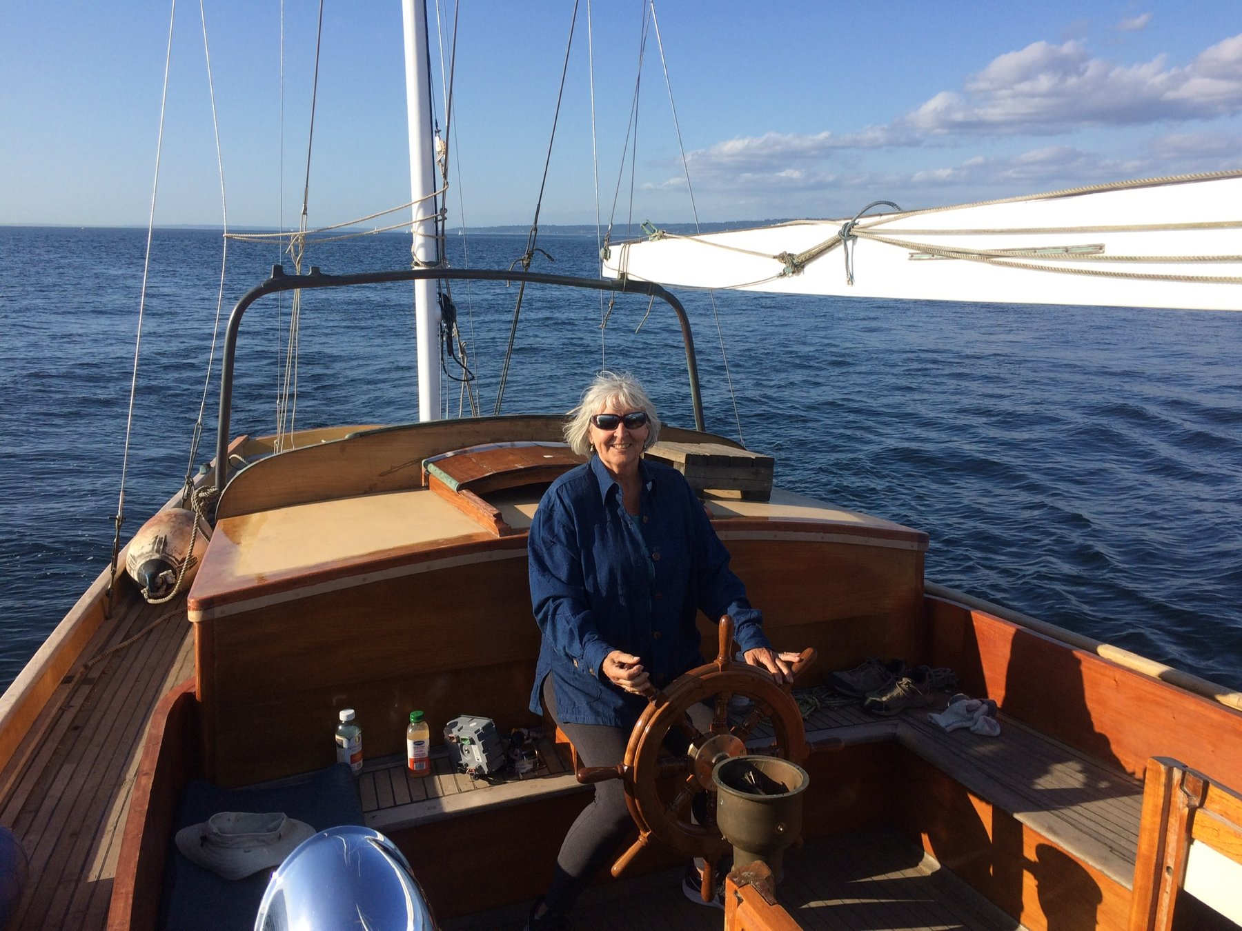 At the helm of the classic 1946 Merry Maiden, a Philip Rhodes design glides through the serene waters of Puget Sound on a perfect summer afternoon, with billowing sails and the Pacific Northwest coastline in the distance.